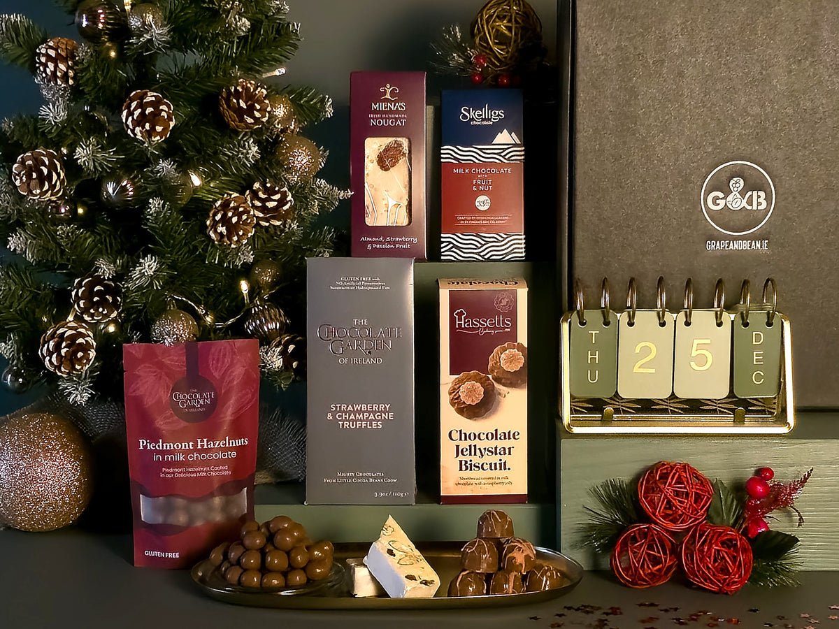 A Christmas-themed gourmet gift hamper featuring an assortment of chocolates, a bag of hazelnuts, and a large gift box, styled with a decorated Christmas tree and festive decorations.