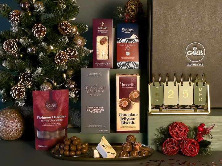 A Christmas-themed gourmet gift hamper featuring an assortment of chocolates, a bag of hazelnuts, and a large gift box, styled with a decorated Christmas tree and festive decorations.