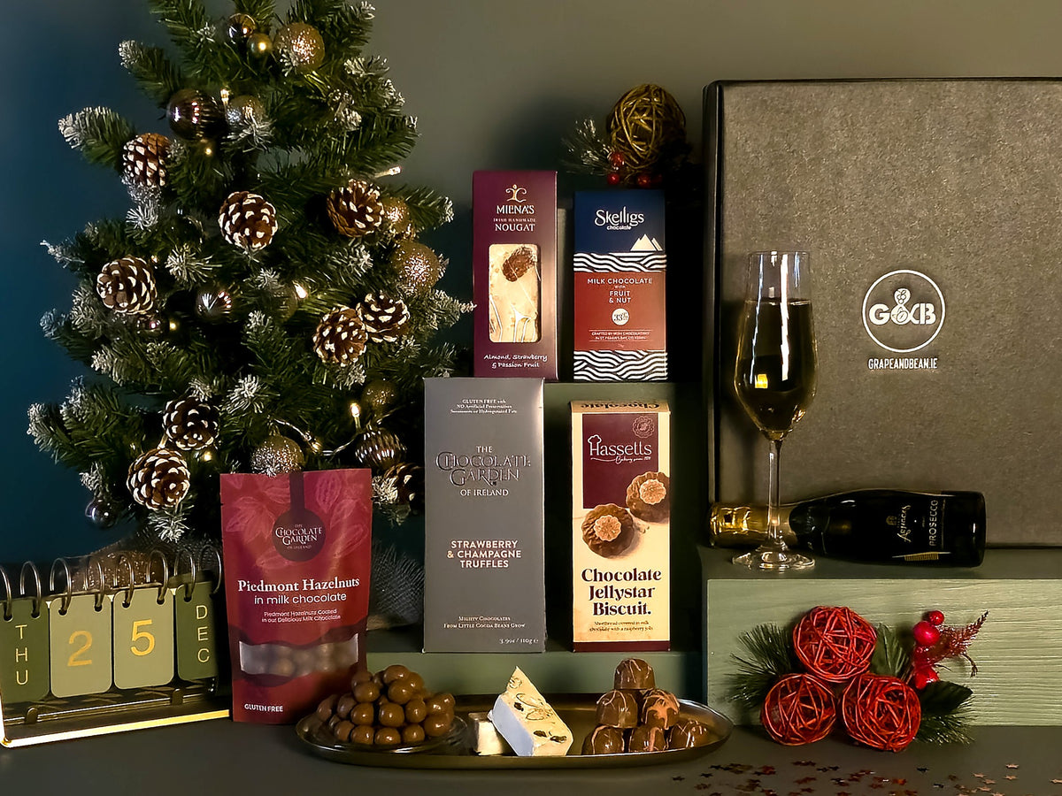 A Christmas-themed gourmet gift set featuring assorted chocolates, a bag of Piedmont hazelnuts, a bottle of prosecco, styled with a decorated Christmas tree, a calendar, and red decorative roses.