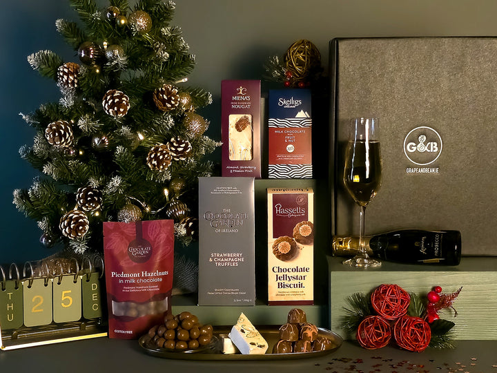 A Christmas-themed gourmet gift set featuring assorted chocolates, a bag of Piedmont hazelnuts, a bottle of prosecco, styled with a decorated Christmas tree, a calendar, and red decorative roses.