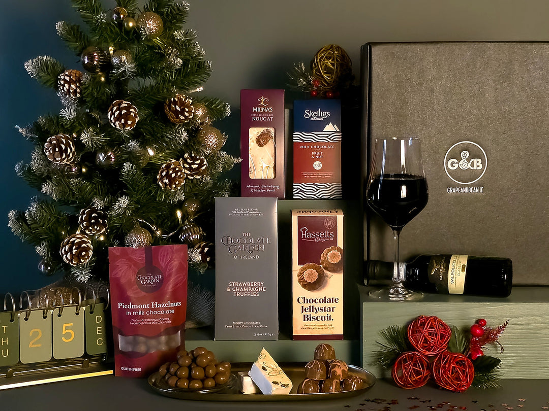 A Christmas-themed gourmet gift hamper featuring an assortment of packaged chocolates, biscuits, and a bag of hazelnuts, arranged alongside a glass of red wine and decorations including a pine tree, a calendar, and red roses.