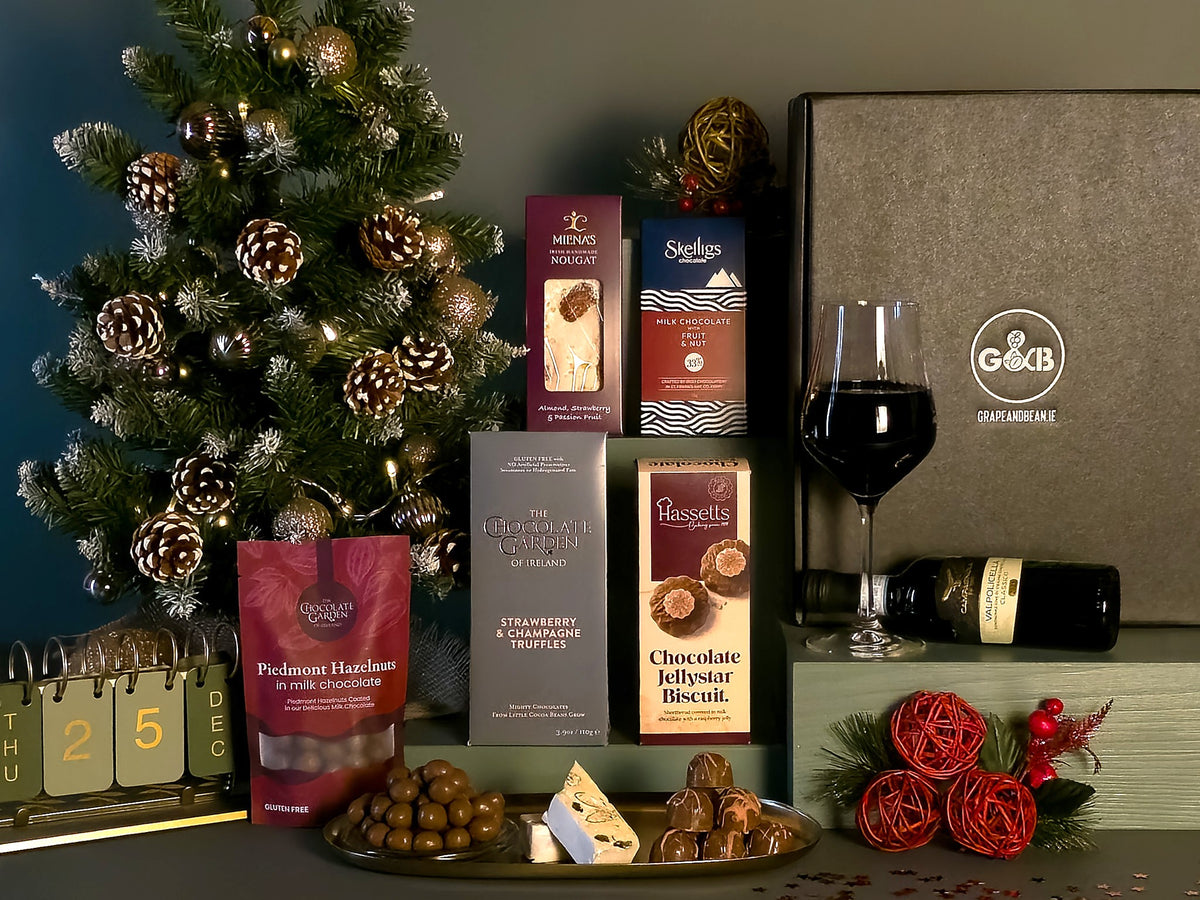 A Christmas-themed gourmet gift hamper featuring an assortment of packaged chocolates, biscuits, and a bag of hazelnuts, arranged alongside a glass of red wine and decorations including a pine tree, a calendar, and red roses.