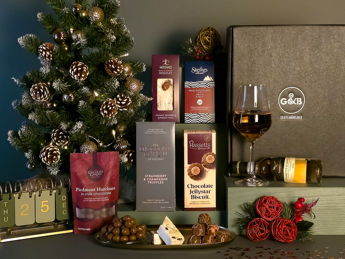 A Christmas-themed gourmet gift hamper featuring an assortment of chocolates, biscuits, and snacks arranged alongside a bottle of wine, a small decorated tree, and a large branded gift box on a dark tabletop.