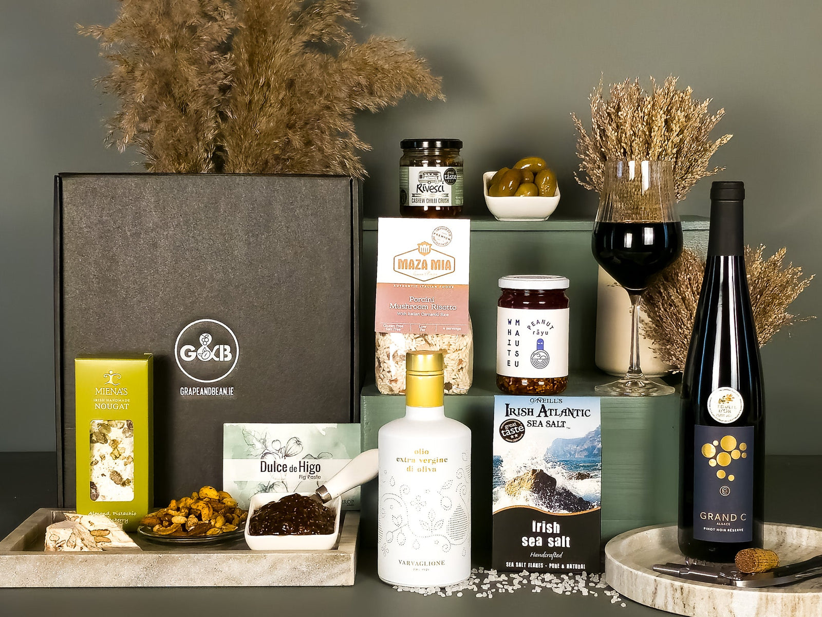 Collection of gourmet items including wine, sea salt, risotto, peanut rayu,fig past and nougat on a gray surface with decorative elements.