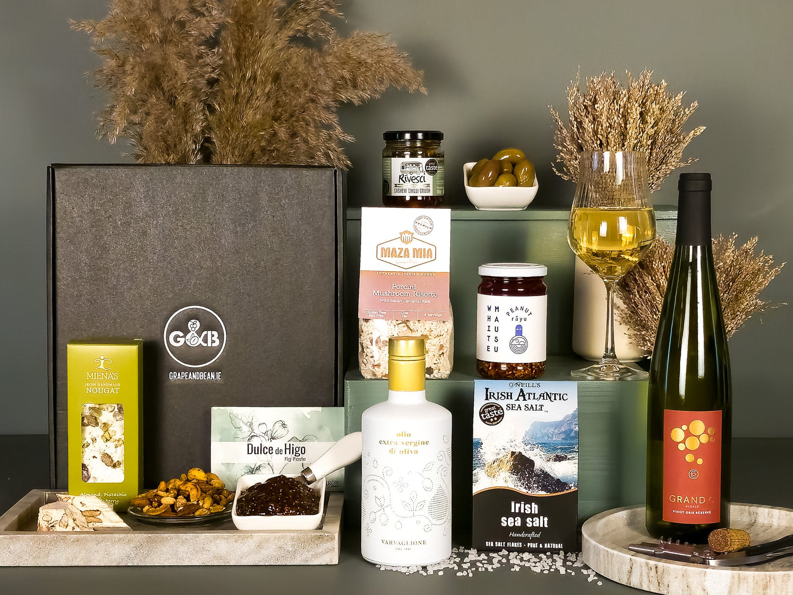 Gourmet gift set with various items including a bottle of Pinot Gris wine, rizotto, peanut rayu, sea salt, olive oil, nougat, fig paste  and snacks on a gray surface