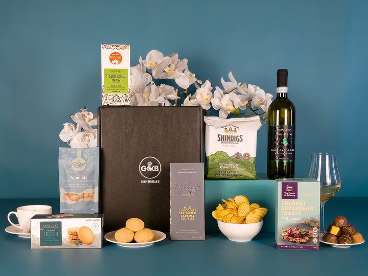 Assorted gourmet items including a bottle of white wine, snacks, biscuits, chocolate, crisps and a black gift box