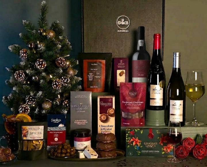 A Christmas-themed gourmet gift hamper display featuring a decorated Christmas tree, bottles of wine, chocolates, spreads, and snacks arranged on a table with glasses, set against a dark background.